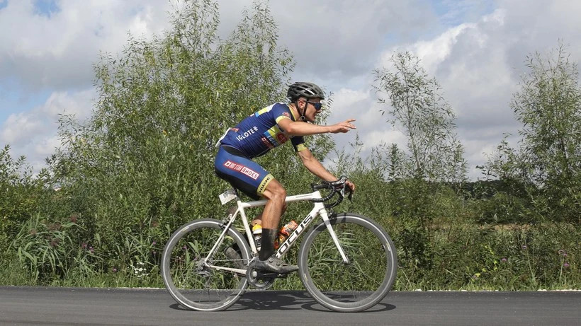 Kolarz w profesjonalnym stroju sportowym jedzie na rowerze szosowym rozciągniętą, asfaltową drogą otoczoną zielenią, unosząc dwie palce w geście pokoju.