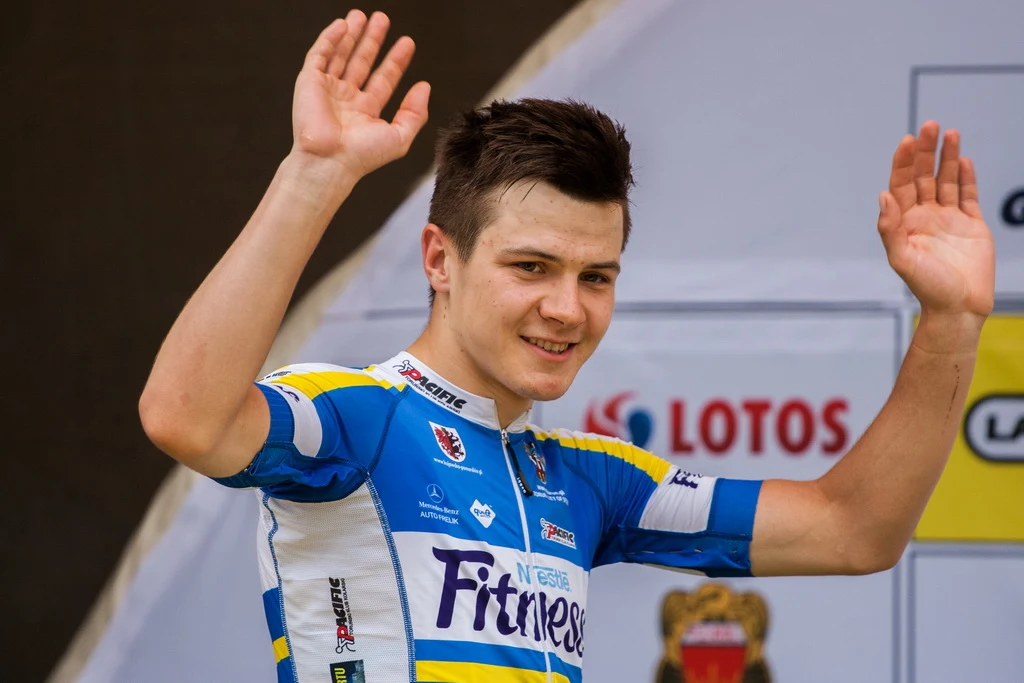 Młody kolarz w sportowej koszulce unosi obie ręce w geście pozdrowienia, na tle planszy z logotypami sponsorów.