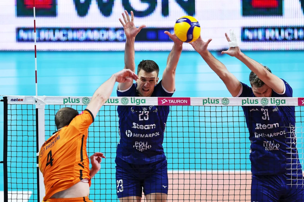 Kadr z meczu Barkom Każany Lwów - Steam Hemarpol Politechnika Częstochowa Dwóch siatkarzy w niebieskich strojach wyskakuje do bloku przy siatce, podczas gdy zawodnik w pomarańczowym stroju atakuje piłkę. Na siatce widoczne są logotypy sponsorów oraz napisy PlusLiga i TAURON, w tle fragment hali sportowej.