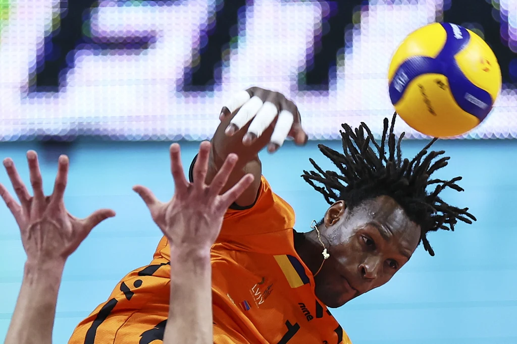 Mousse Gueye, środkowy Barkomu Każany Lwó Sportowiec w pomarańczowej koszulce wykonuje dynamiczny atak w trakcie meczu siatkówki, tuż nad siatką widać piłkę oraz dłonie obrońcy próbującego zablokować atak.