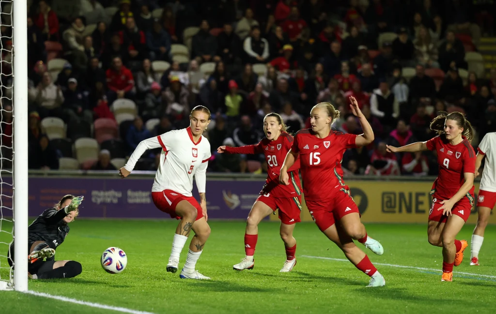 Kadr z meczu Polska - Walia Zawodniczki dwóch drużyn piłkarskich rywalizują o piłkę podczas meczu, jedna z nich stoi blisko bramki przeciwników, a bramkarka próbuje obronić strzał. Na trybunach widownia obserwuje rozgrywkę.