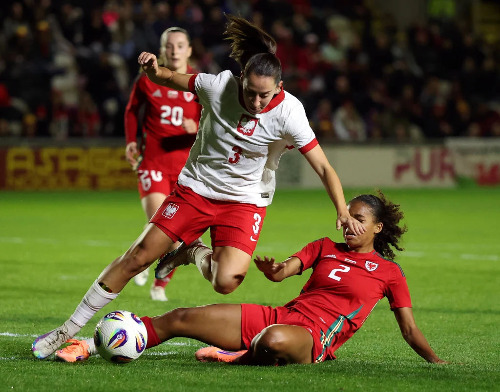 Kadr z meczu Polska - Walia Dwie piłkarki w trakcie zaciętej walki o piłkę podczas meczu piłki nożnej kobiet, jedna z nich próbuje przejąć piłkę wślizgiem, obie skupione na grze, w tle widać kolejne zawodniczki i fragment stadionu.