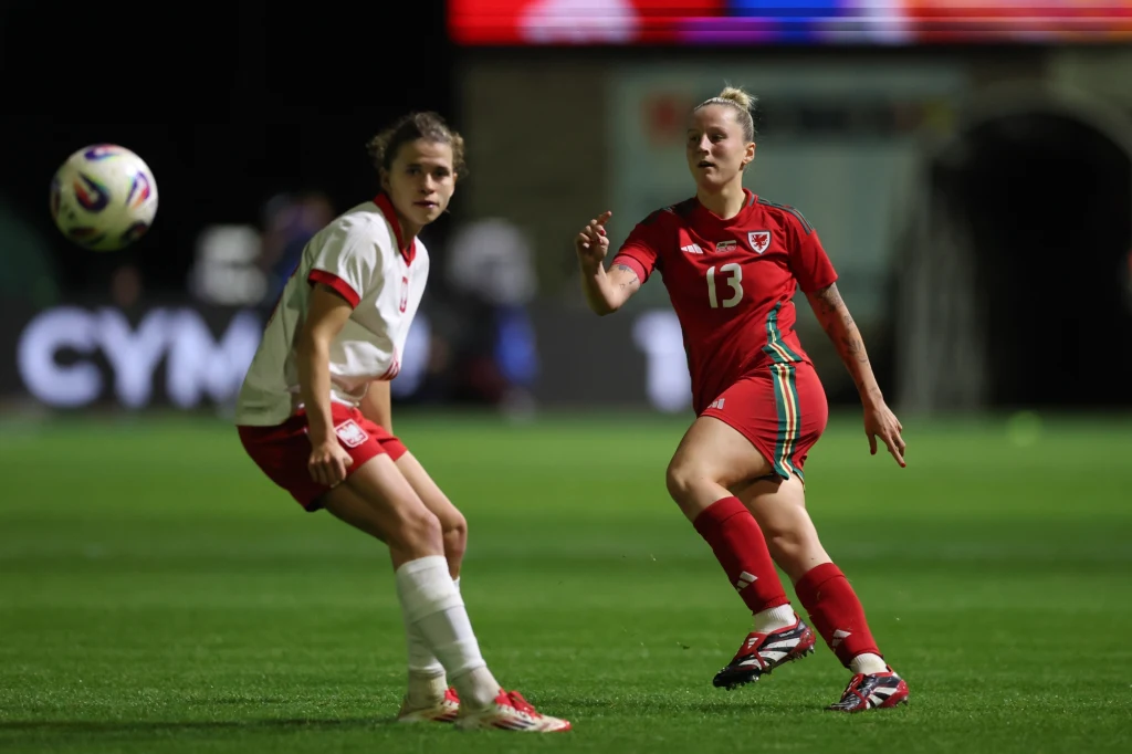 Kadr z meczu Polska - Walia Dwie piłkarki w trakcie meczu, jedna ubrana w biały strój sportowy, druga w czerwony z numerem 13. Kobiety skupione są na grze, w tle widoczne rozmyte światła stadionu.