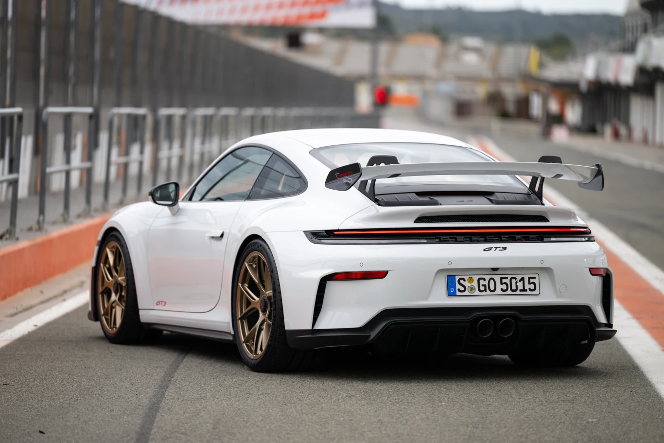 Biały sportowy samochód Porsche 911 GT3 z dużym tylnym spojlerem, złotymi felgami oraz niemiecką tablicą rejestracyjną stoi na torze wyścigowym, otoczony barierkami i pustymi trybunami w tle.
