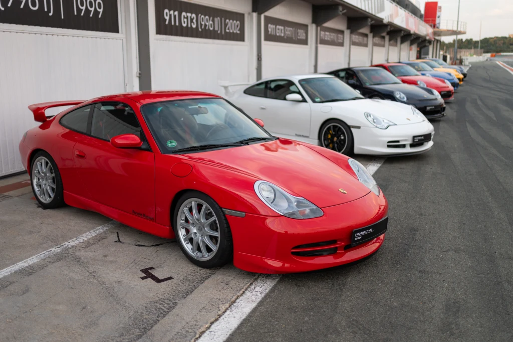Rząd sportowych samochodów Porsche zaparkowanych przy torze wyścigowym, z przodu dominuje czerwone auto, a w tle widać kolejne modele w różnych kolorach.