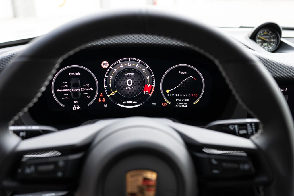 Cyfrowe liczniki na desce rozdzielczej sportowego samochodu Porsche, wyraźnie widoczny prędkościomierz, wskaźnik mocy oraz szczegóły dotyczące ogumienia, kierownica z logo Porsche na pierwszym planie.