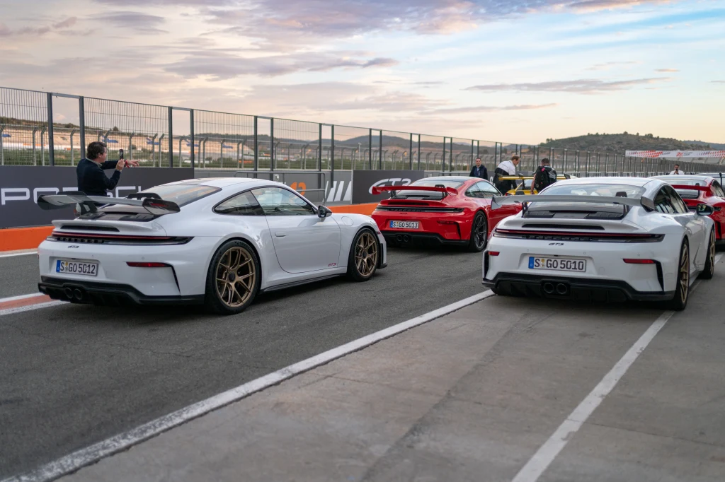 Kilka sportowych samochodów Porsche w jasnych kolorach ustawionych na torze wyścigowym, w tle osoby fotografujące lub przygotowujące auta, metalowe ogrodzenie oraz widoczne niebo z lekkim zachmurzeniem.