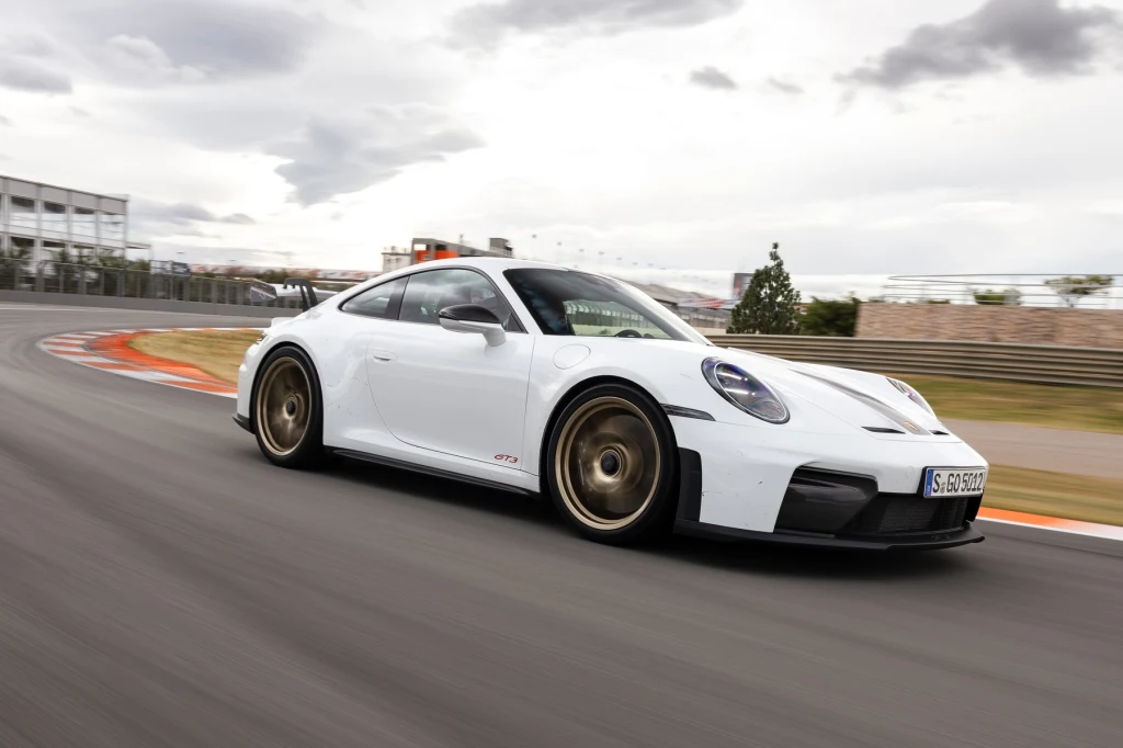 Biały samochód sportowy Porsche 911 GT3 poruszający się po zakręcie toru wyścigowego, w tle widoczne ogrodzenie i budynki torowe, dynamiczna scena z wyraźnym ruchem pojazdu.