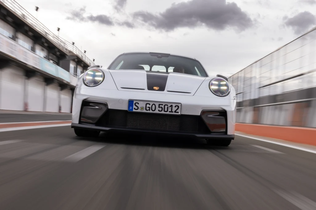 Nowoczesny biały samochód sportowy marki Porsche poruszający się po torze wyścigowym, ujęcie z perspektywy niskiej, podkreślające dynamikę oraz agresywną stylistykę pojazdu. W tle widać trybuny i barierki toru, a na niebie chmury.