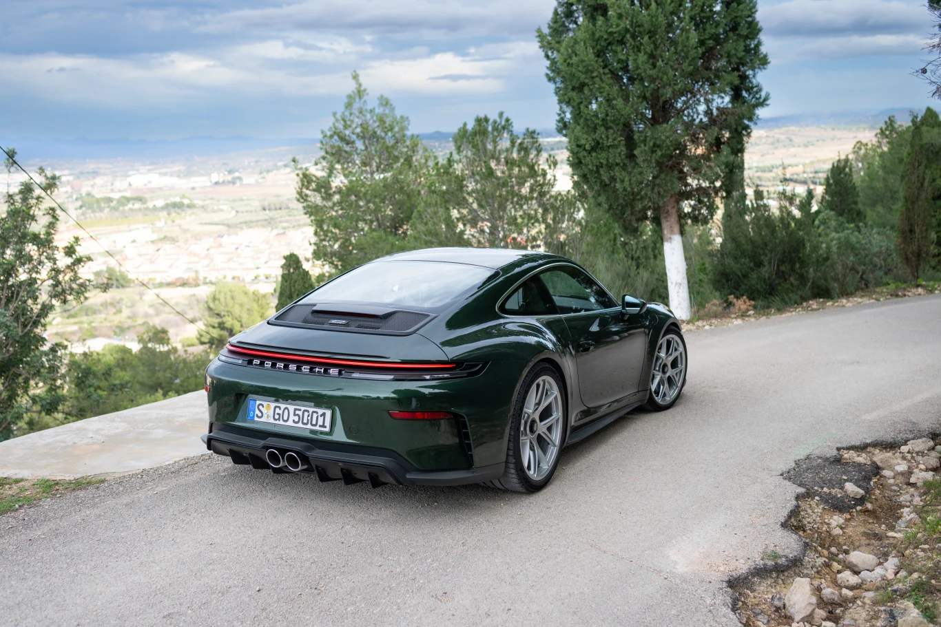 Zielony sportowy samochód Porsche zaparkowany na wąskiej asfaltowej drodze, otoczonej drzewami i z widokiem na dolinę oraz rozciągającą się panoramę miasta w oddali.