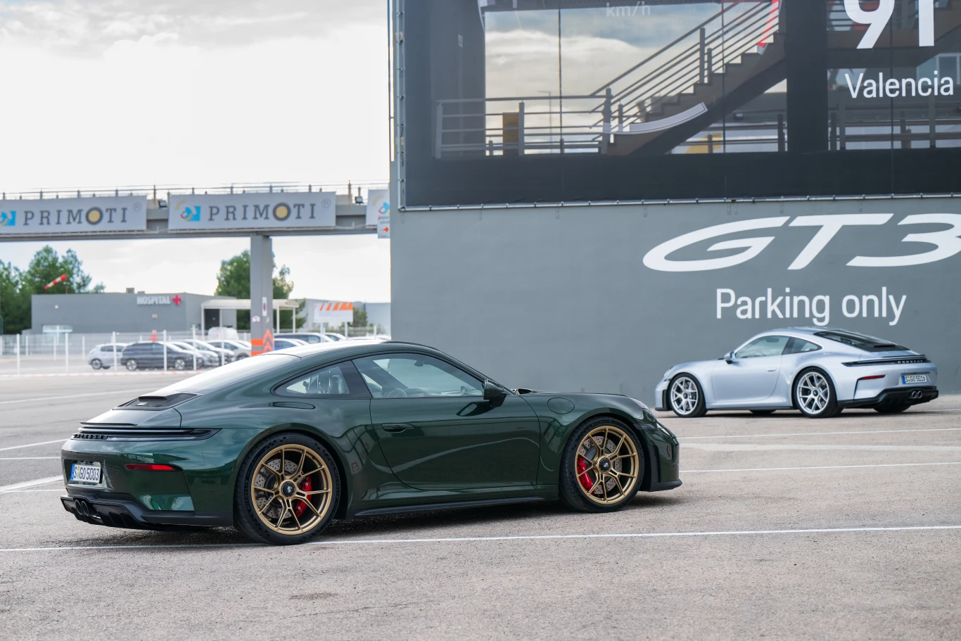 Dwa samochody sportowe Porsche GT3 zaparkowane obok toru wyścigowego, jeden w kolorze ciemnozielonym z felgami w kolorze złotym, drugi srebrny. W tle duży napis GT3 oraz oznaczenie miejsca parkingowego dedykowanego tym modelom.