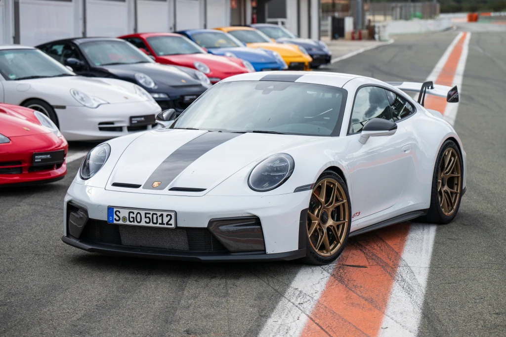 Białe sportowe auto Porsche 911 GT3 z czarnym paskiem na masce i złotymi felgami stoi zaparkowane na torze wyścigowym, w tle szereg kolorowych samochodów sportowych.