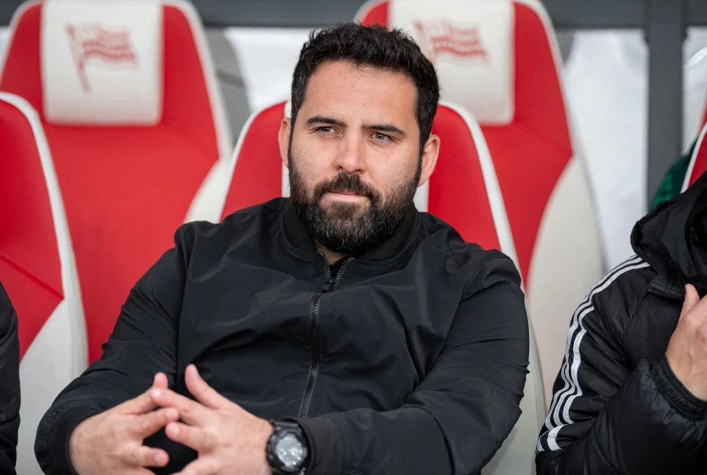 Mężczyzna z brodą w czarnym ubraniu siedzi na stadionowej ławce z czerwono-białymi siedzeniami, trzymając splecione dłonie, obok niego widoczne fragmenty innych osób.