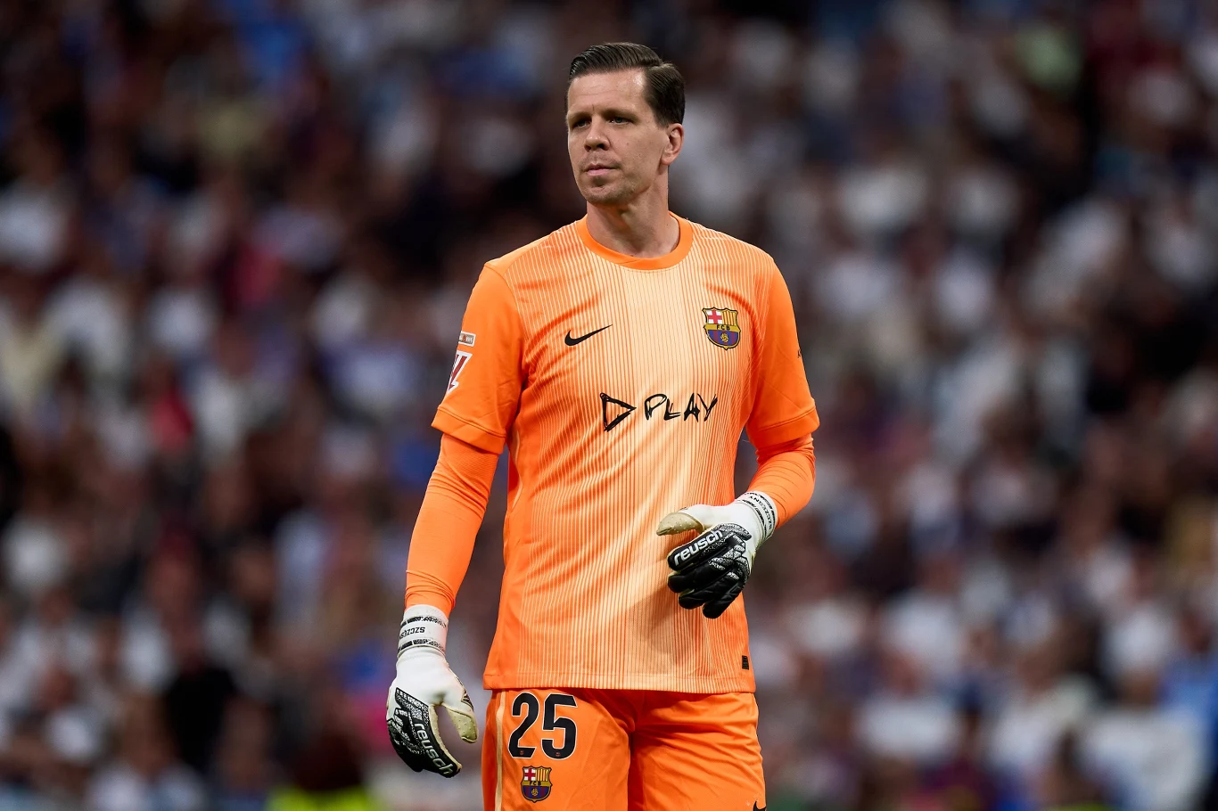 Wojciech Szczęsny Bramkarz w pomarańczowym stroju sportowym z numerem 25 i herbem klubu piłkarskiego na piersi stoi na tle widowni stadionu trzymając piłkę w jednej ręce.