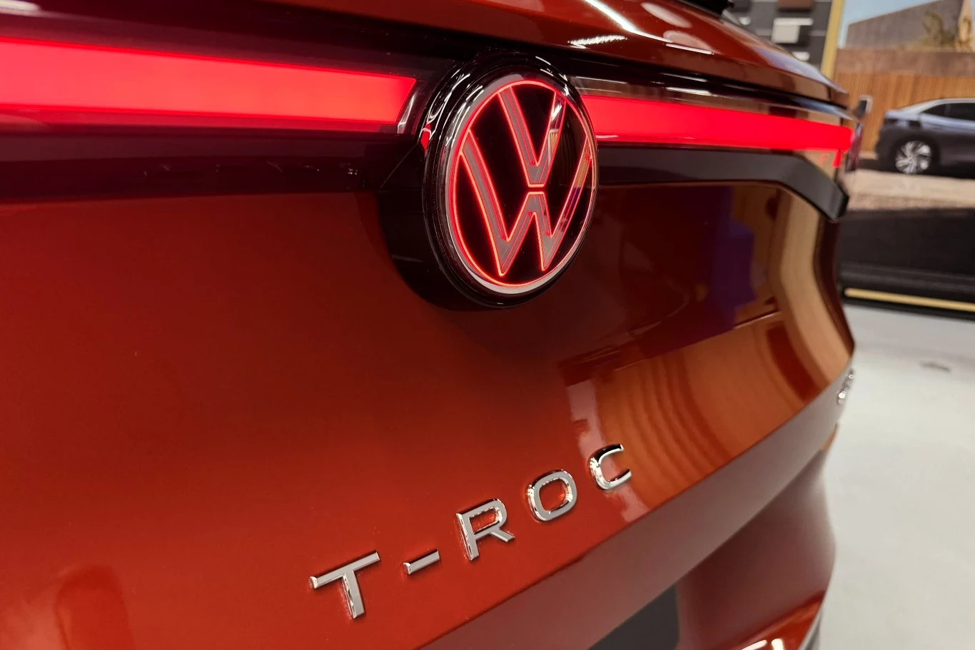 Czerwony tył samochodu Volkswagen T-Roc z podświetlonym logo VW oraz charakterystycznym napisem T-ROC, widoczne detale reflektora i fragment otoczenia w tle.