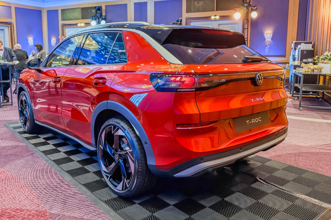 Czerwony Volkswagen T-Roc zaparkowany na czarnej macie ekspozycyjnej w pomieszczeniu o eleganckim wystroju, z dobrze widocznymi refleksami światła na karoserii i elementami wystawowymi w tle.