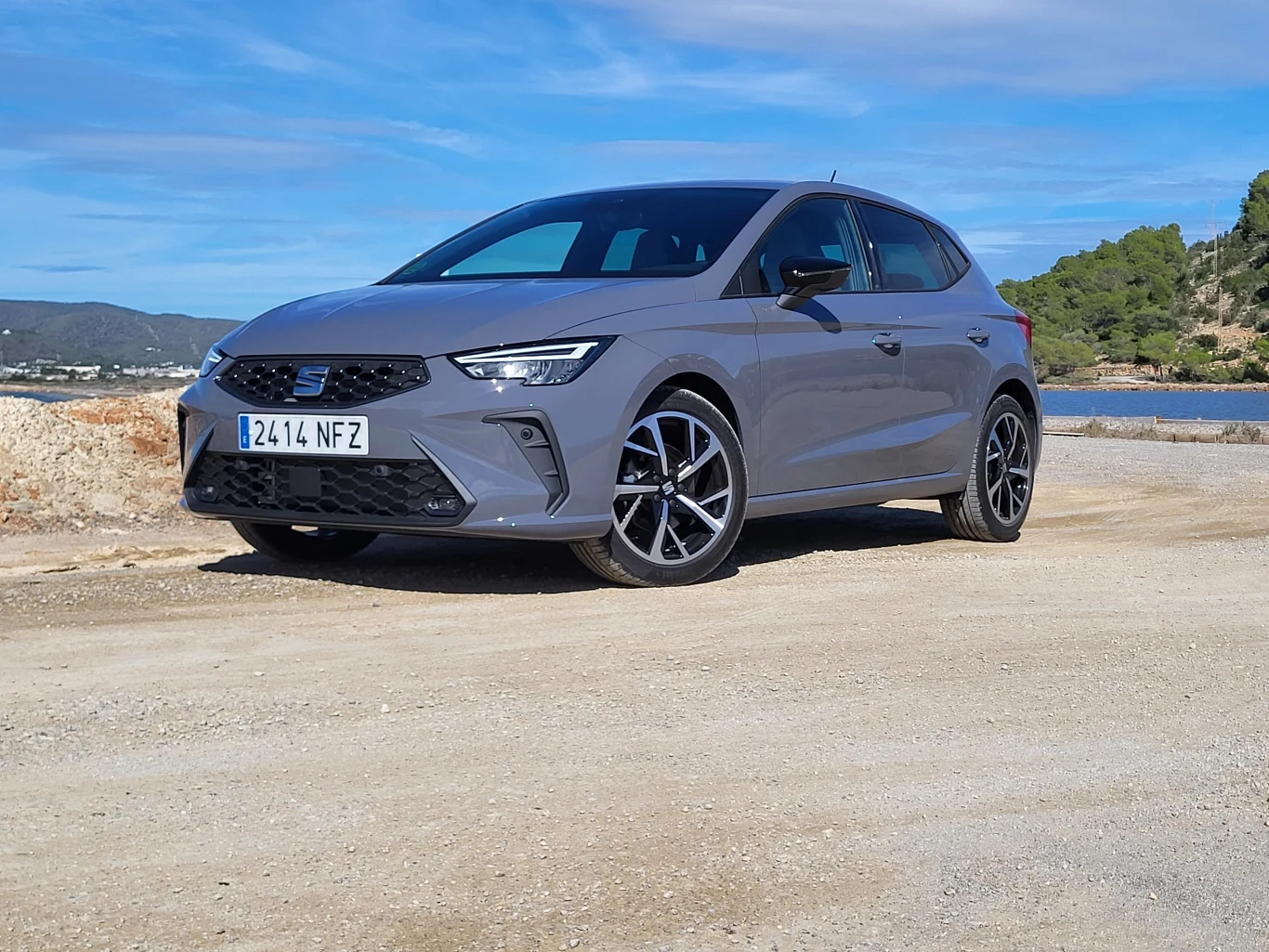 Nowoczesny, szary hatchback marki Seat zaparkowany na żwirowym podłożu w słonecznym, nadmorskim krajobrazie. Samochód wyróżnia się sportowymi felgami i designerskimi reflektorami, w tle widoczne wzgórza i zieleń.