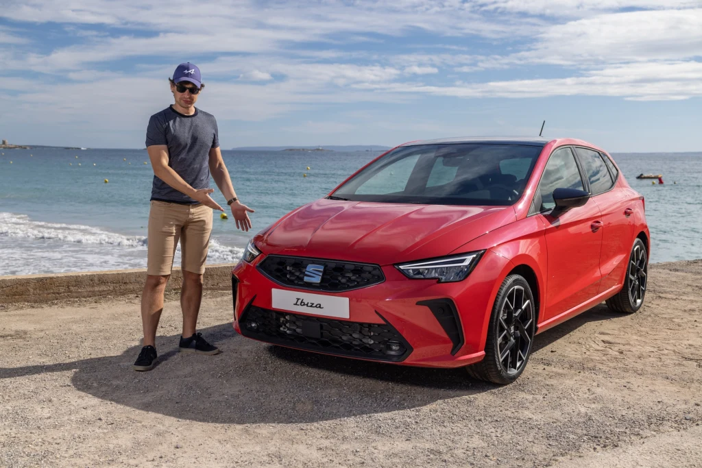 Długość nadwozi Ibizy i Arony wydłużyła się symbolicznie - o, odpowiednio, 1,1 i 1 cm. To rezultat zastosowania nowego zderzaka. Mężczyzna w okularach przeciwsłonecznych i czapce prezentuje czerwony samochód marki Seat Ibiza zaparkowany nad morzem, w tle błękitna woda i lekko pochmurne niebo.