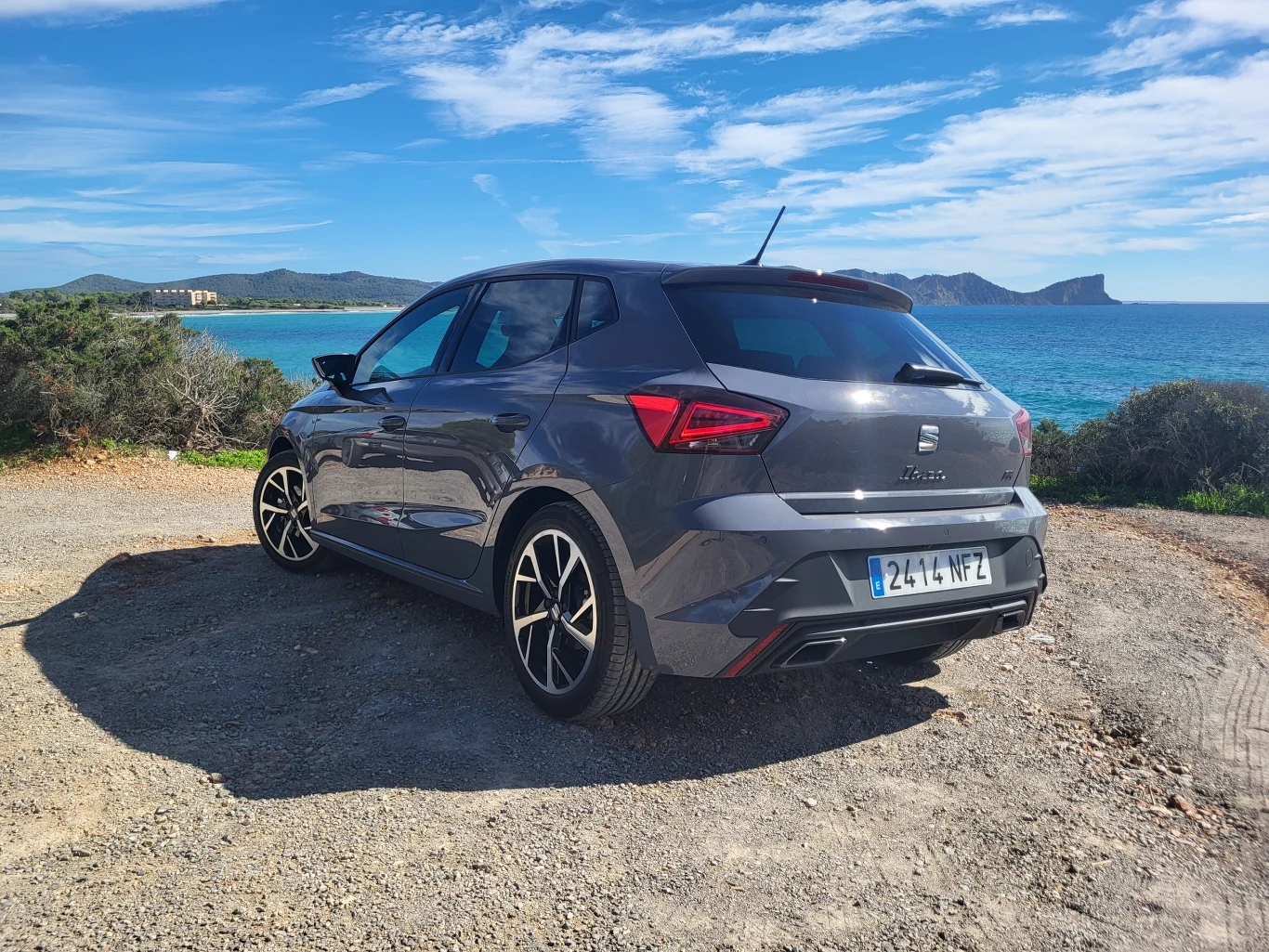 Szary samochód osobowy marki Seat zaparkowany na żwirowym placu, w tle widoczne turkusowe morze, pagórkowate wybrzeże oraz niebieskie niebo z lekkim zachmurzeniem.