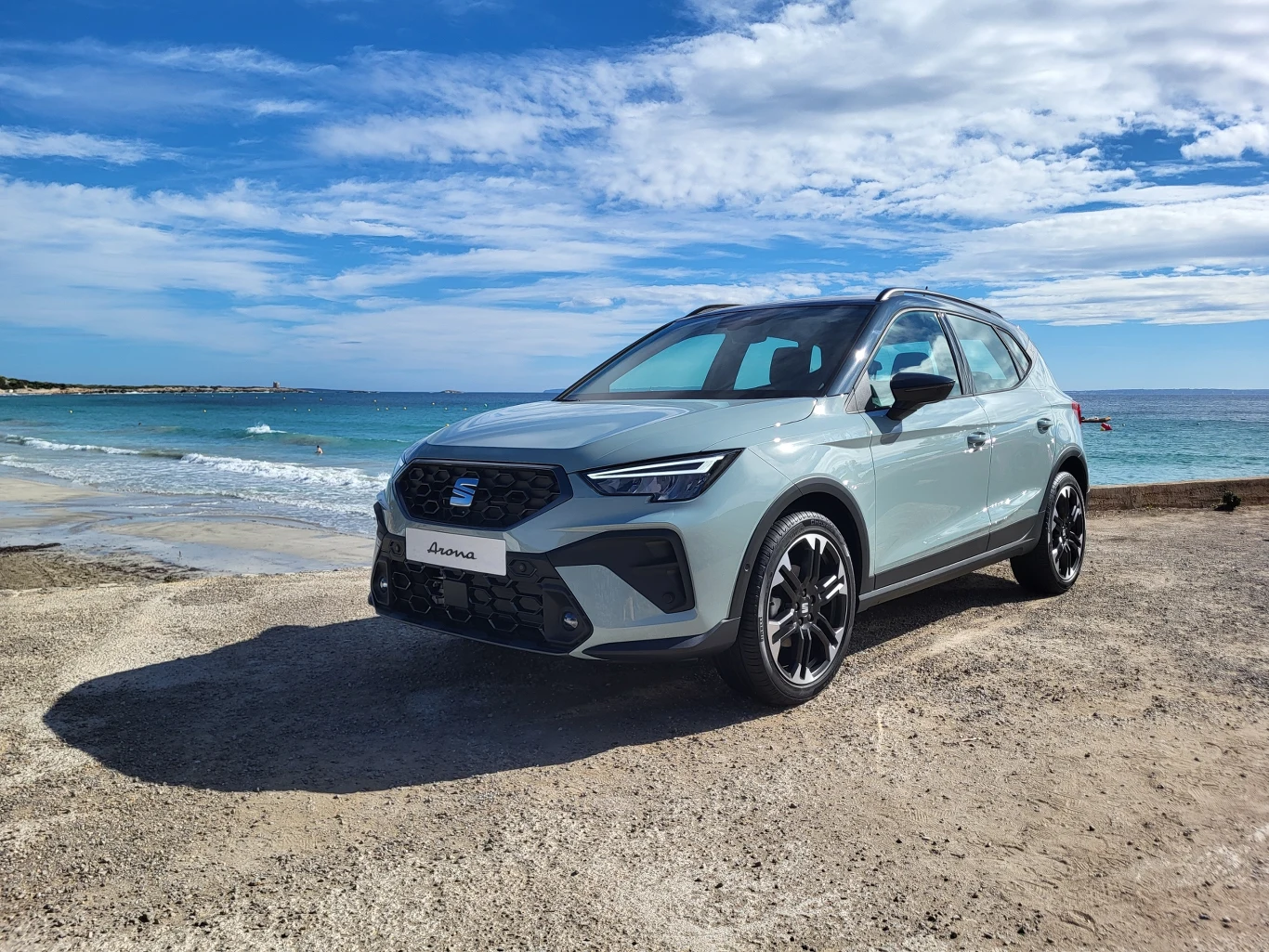 Nowoczesny srebrny SUV marki SEAT Arona zaparkowany na plaży z widokiem na morze, niebo pokryte częściowo chmurami, fale delikatnie uderzają o brzeg.