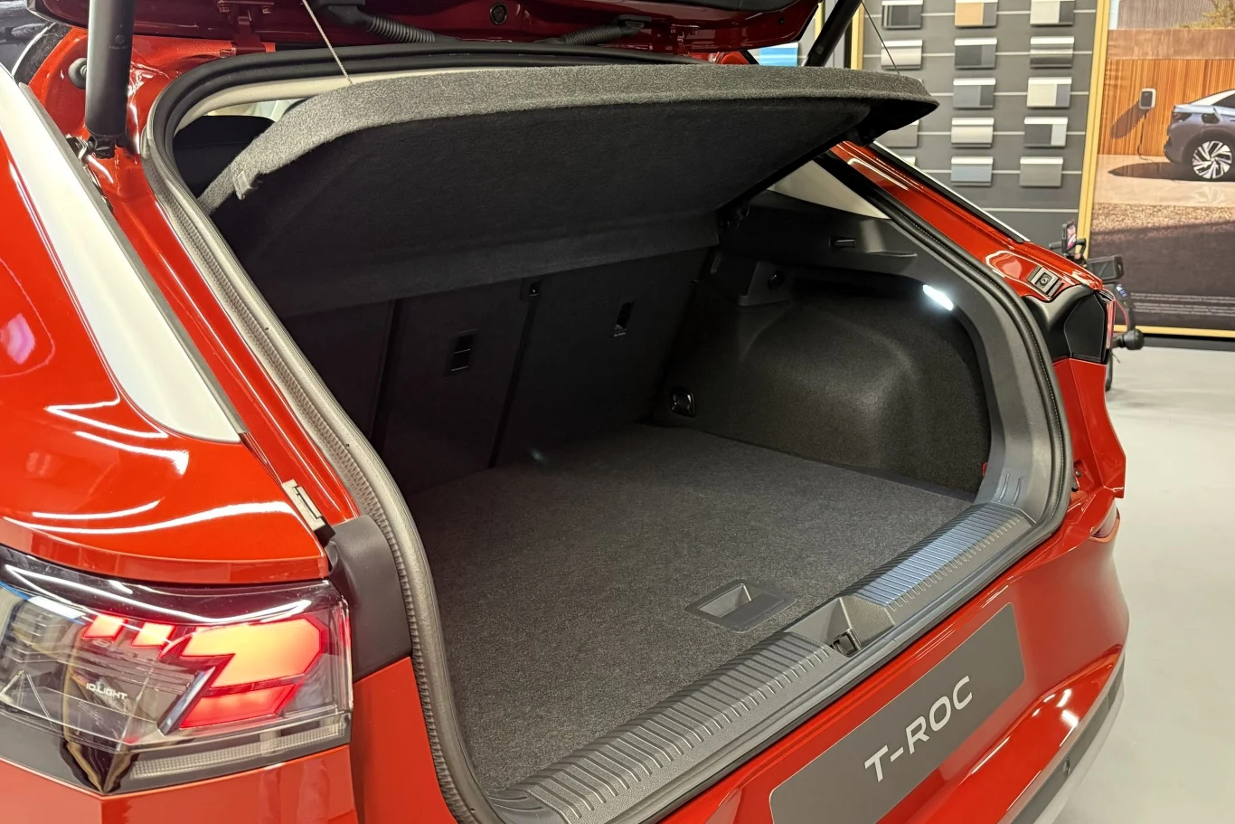 Otwarty bagażnik czerwonego samochodu marki Volkswagen T-Roc, widoczna pusta przestrzeń bagażowa, tapicerka w ciemnoszarym kolorze, otwarte tylne drzwi, w tle ekspozycja z elementami wykończenia wnętrz.
