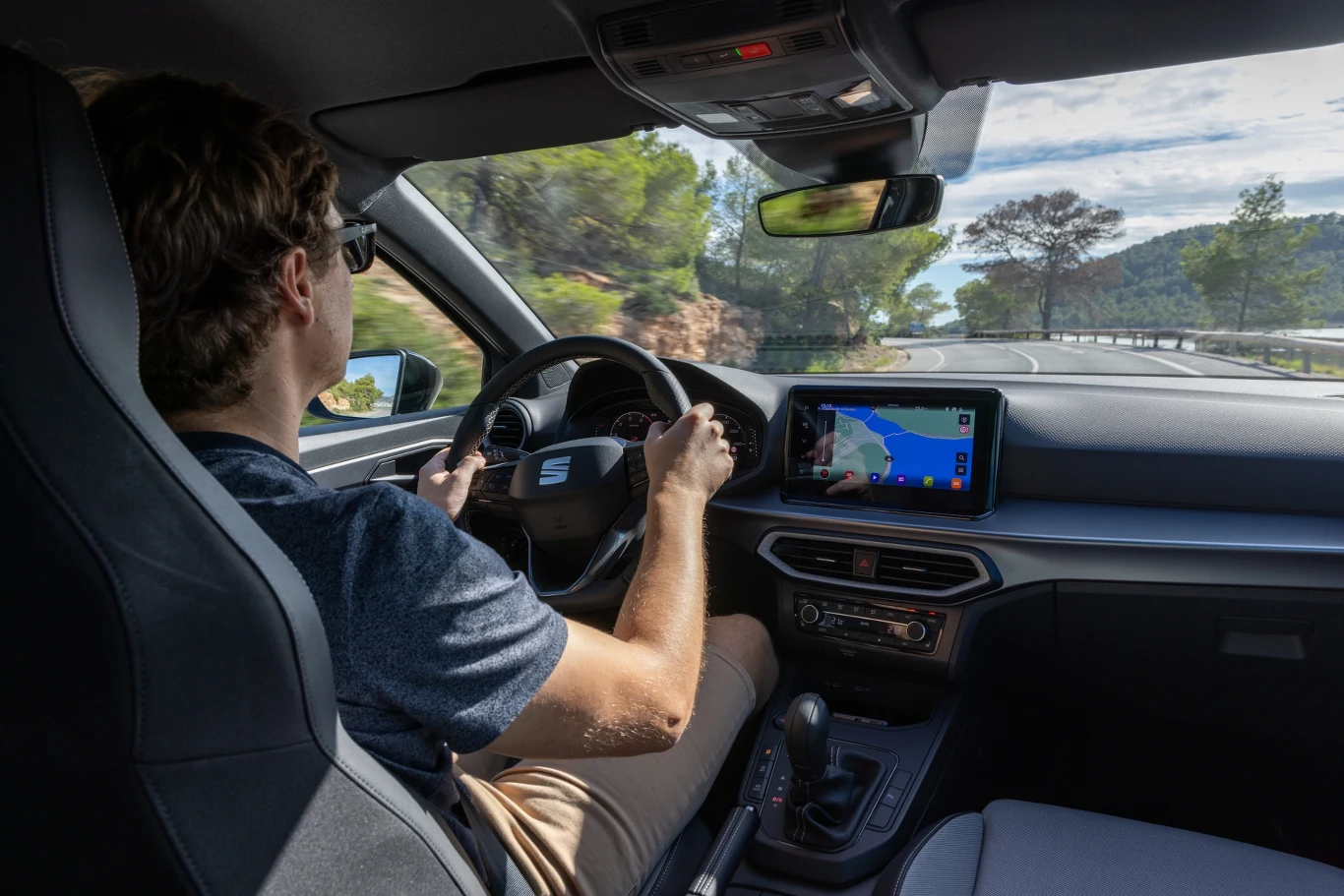 Mężczyzna prowadzi samochód, korzystając z nawigacji GPS na pokładzie, jasne wnętrze auta i widok na krętą, otoczoną zielenią drogę za oknem.