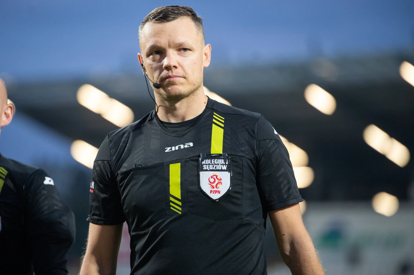 Damian Krumplewski Sędzia piłkarski w czarnym stroju z logo PZPN oraz słuchawką na uchu, stojący na tle rozjaśnionego stadionu.