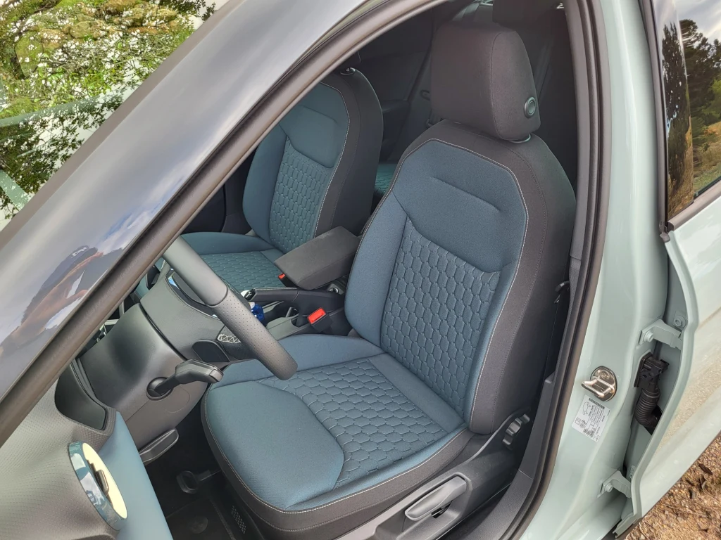 Za to na fotelach pojawiła się nowa, tłoczona tkanina - wyraźnie lepszej jakości niż dotąd. Wnętrze nowoczesnego samochodu z dwoma przednimi fotelami pokrytymi niebieską tapicerką z dekoracyjnym przeszyciem, widoczna kierownica, konsola centralna oraz otwarte drzwi z fragmentem szyby.