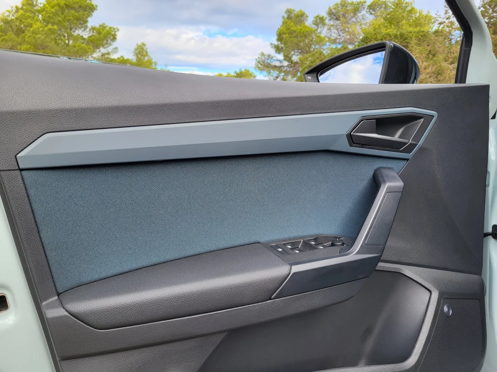 Wykończenie materiałowe drzwi może nawiązywać do tapicerki na fotelach - kolorowe wstawki wyraźnie ożywiają wnętrze Arony. Wnętrze bocznej części drzwi samochodu z nowoczesnym designem, wykończeniem z materiału tekstylnego i tworzywa sztucznego oraz panel sterowania szyby po stronie kierowcy, w tle fragment lusterka bocznego, drzewa i niebo.