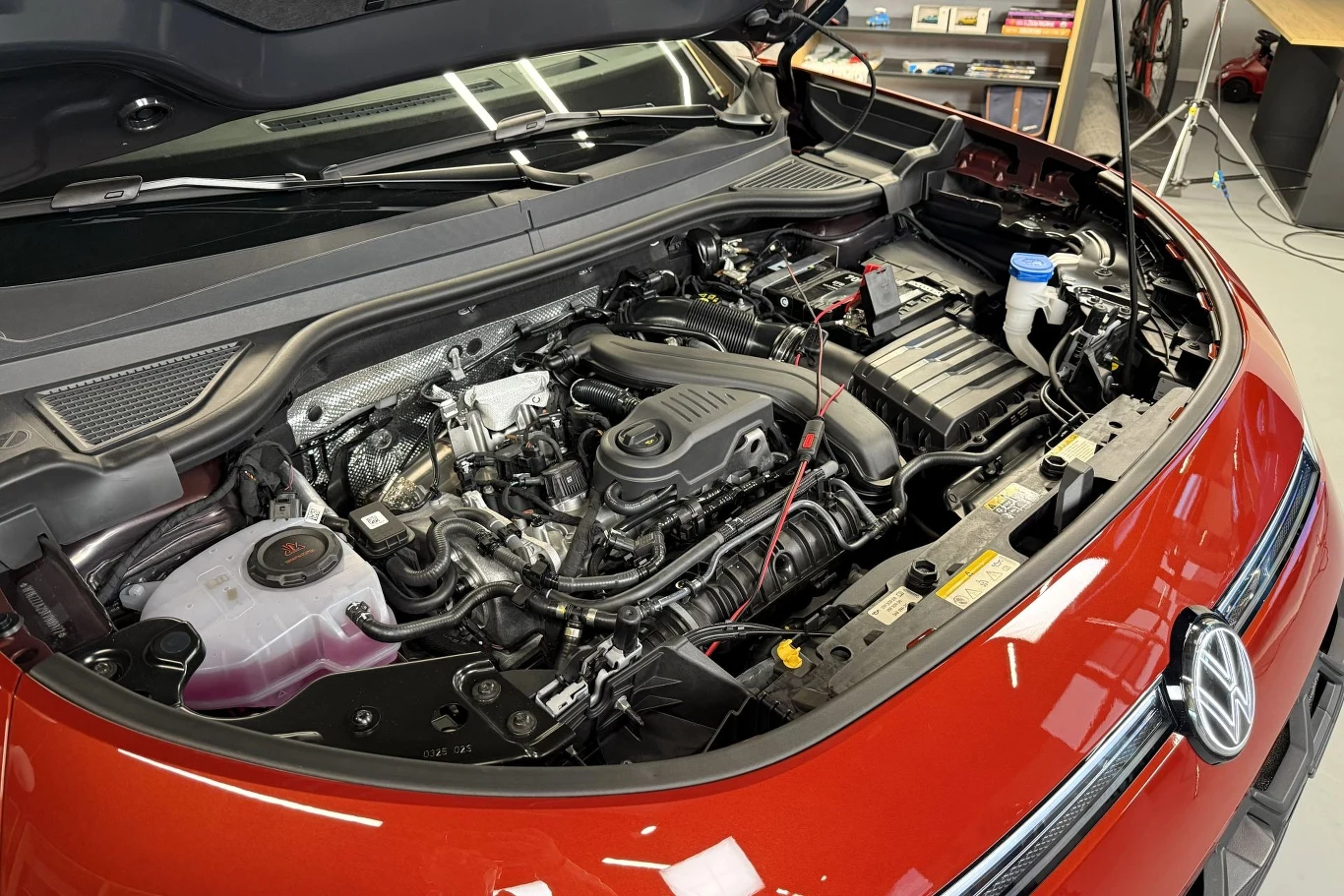 Otwarta maska czerwonego samochodu marki Volkswagen, pod którą widoczny jest silnik wraz z elementami mechanicznymi i przewodami w warsztatowym otoczeniu.