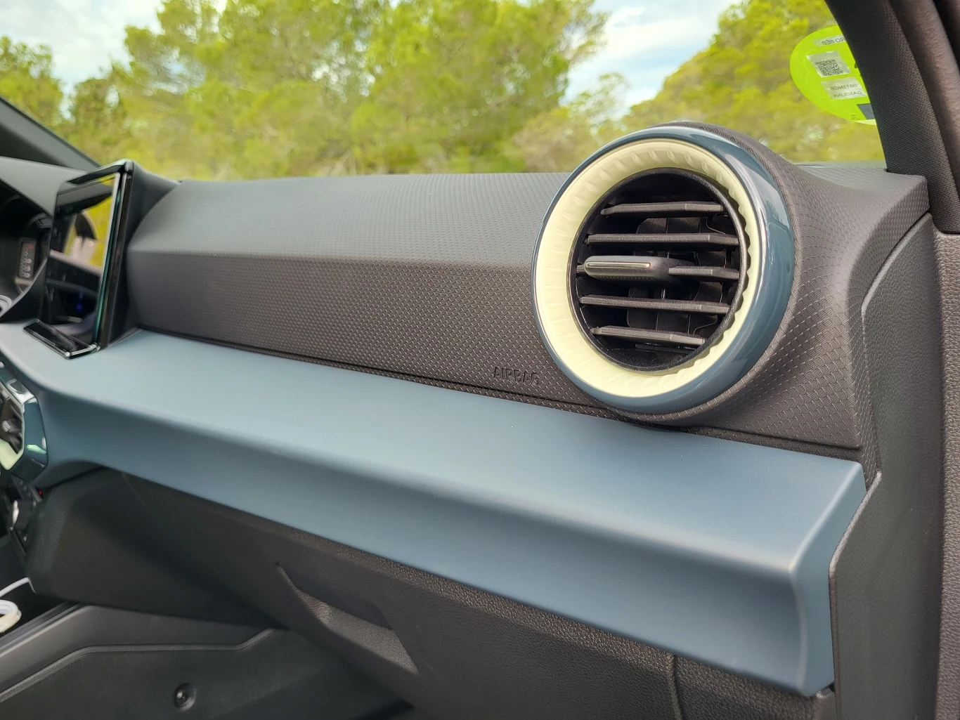 Deska rozdzielcza samochodu z wyraźnie widocznym okrągłym nawiewem powietrza, kolorem dominującym jest czarny i niebieski, w tle widoczne rozmyte drzewa.