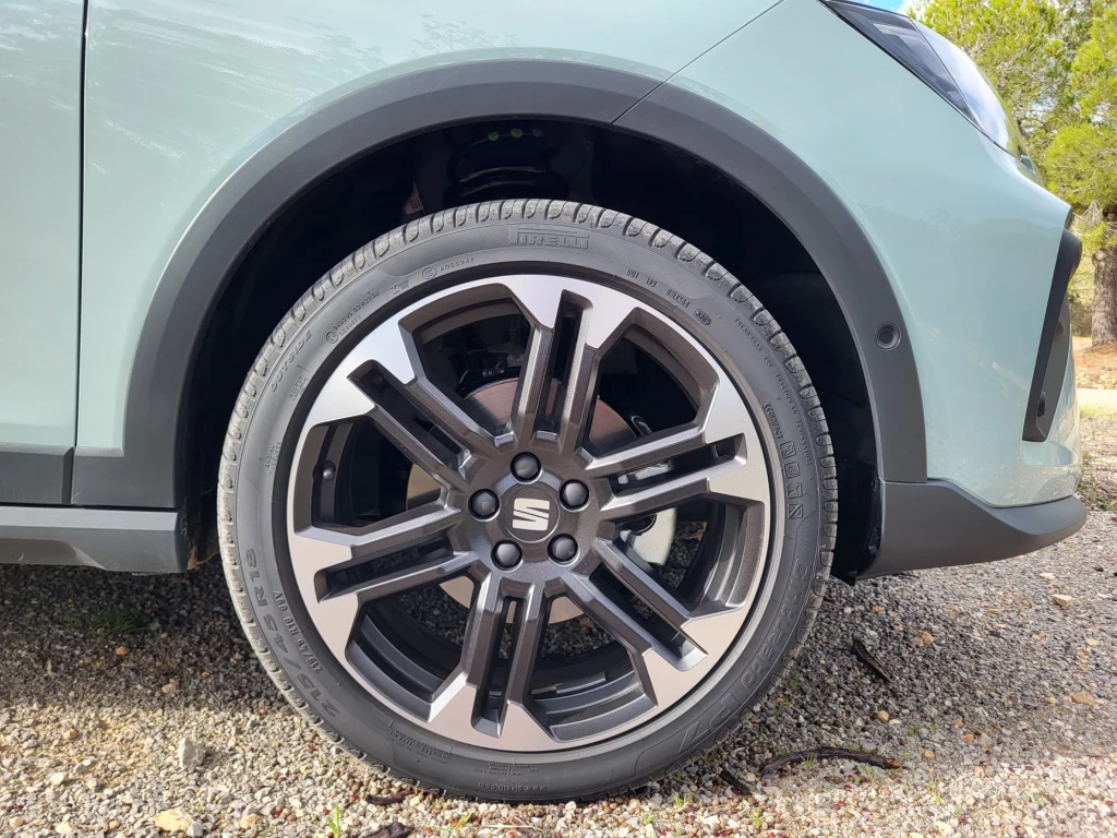 Obręcze Arony (na zdjęciu) mogą mieć rozmiar od 16 do 18 cali, w Ibizie dostępne są także 15-calowe. Fragment samochodu z wyeksponowanym nowoczesnym kołem oraz felgą aluminiową na tle żwirowego podłoża, widoczny jest kawałek nadwozia oraz przednie reflektory.