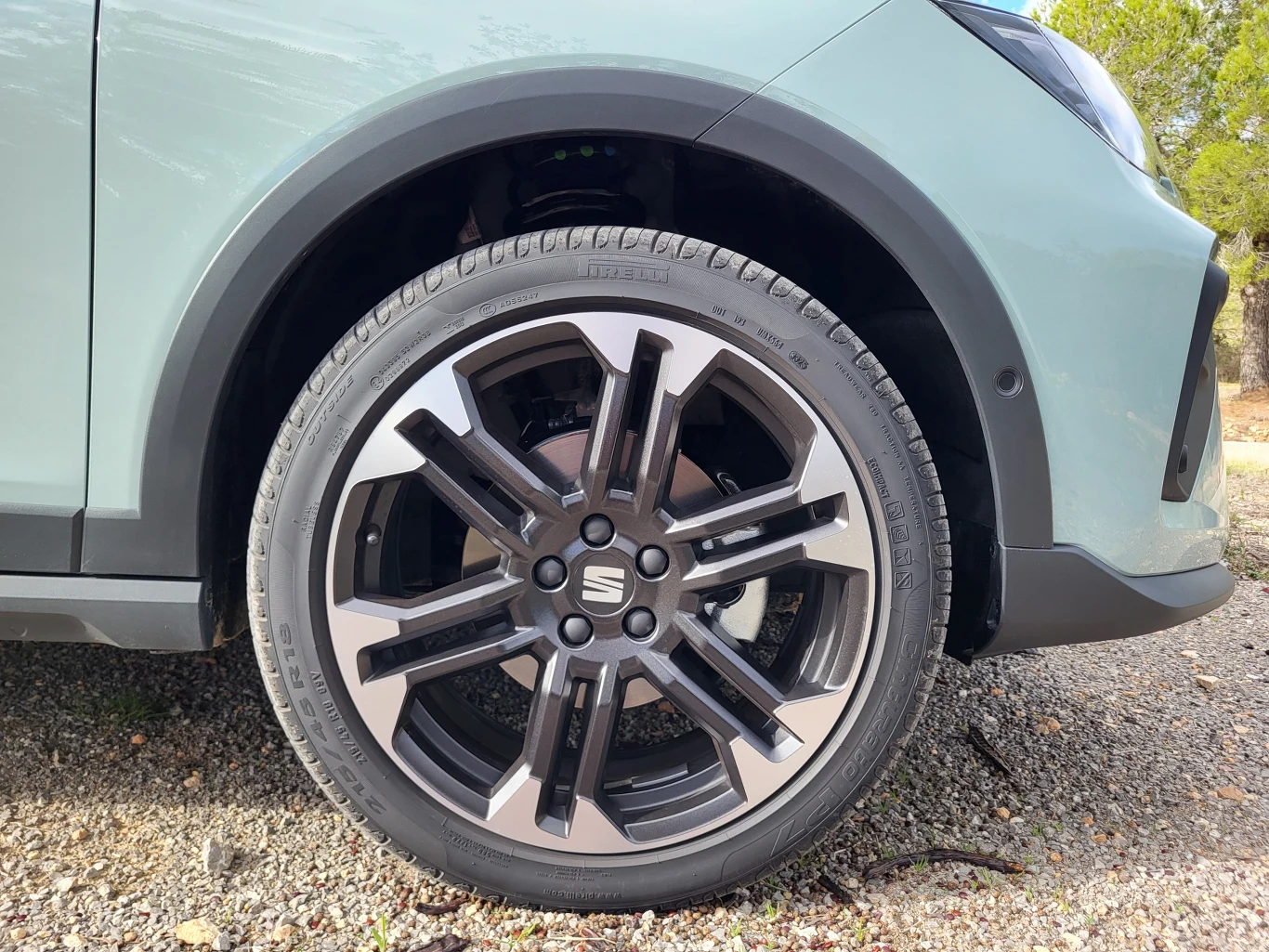 Fragment samochodu z wyeksponowanym nowoczesnym kołem oraz felgą aluminiową na tle żwirowego podłoża, widoczny jest kawałek nadwozia oraz przednie reflektory.