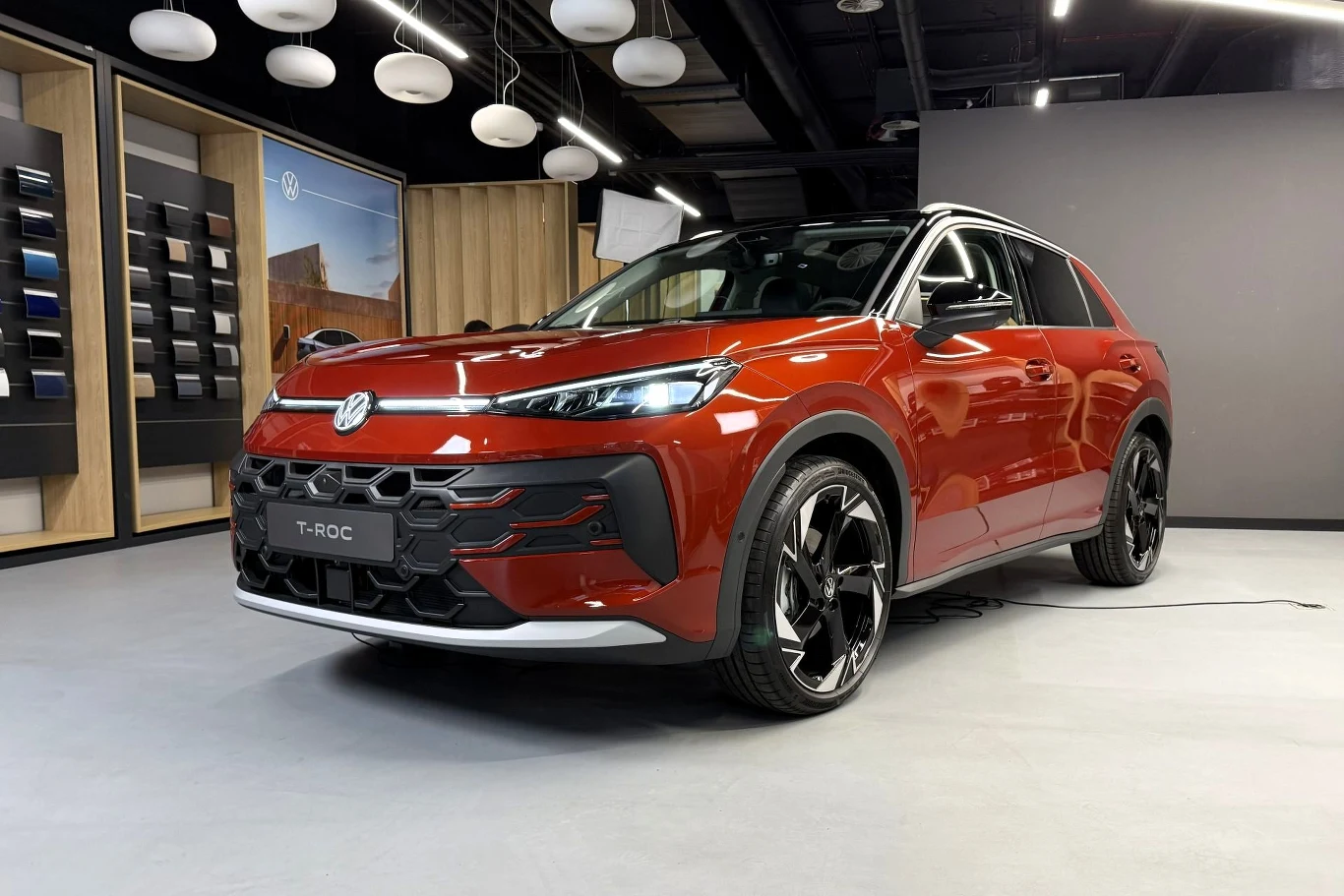 Czerwony samochód Volkswagen T-Roc w nowoczesnym salonie, otoczony eleganckim wnętrzem z designerskimi lampami oraz panelami prezentującymi różne opcje wykończenia pojazdu.