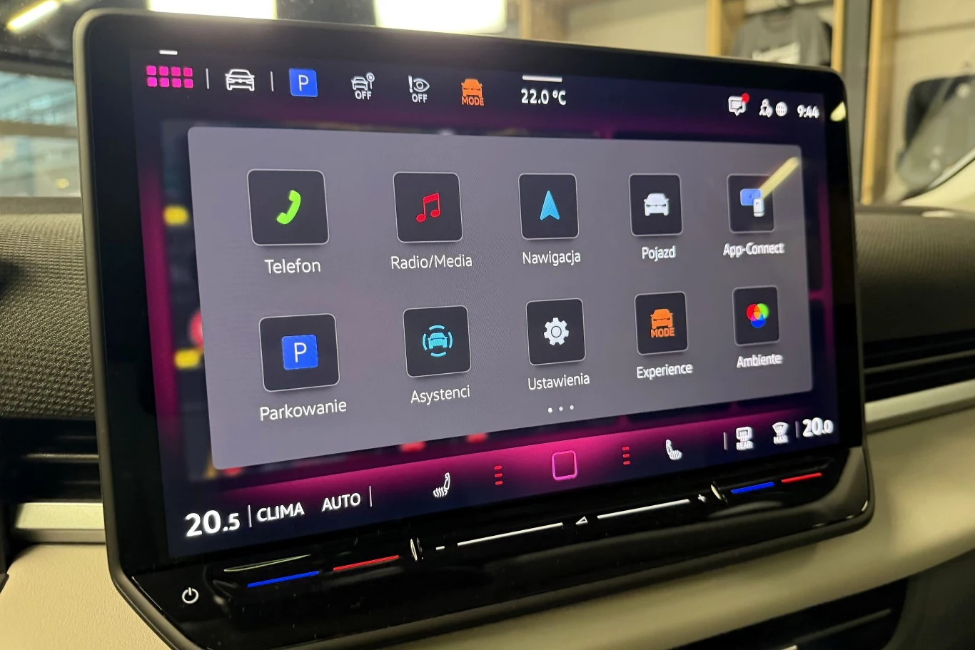 Dotykowy ekran systemu multimedialnego w nowoczesnym samochodzie wyświetlający menu główne z ikonami do nawigacji, sterowania telefonem, radiem, ustawieniami pojazdu i innymi funkcjami. Otoczenie wnętrza auta widoczne w tle.