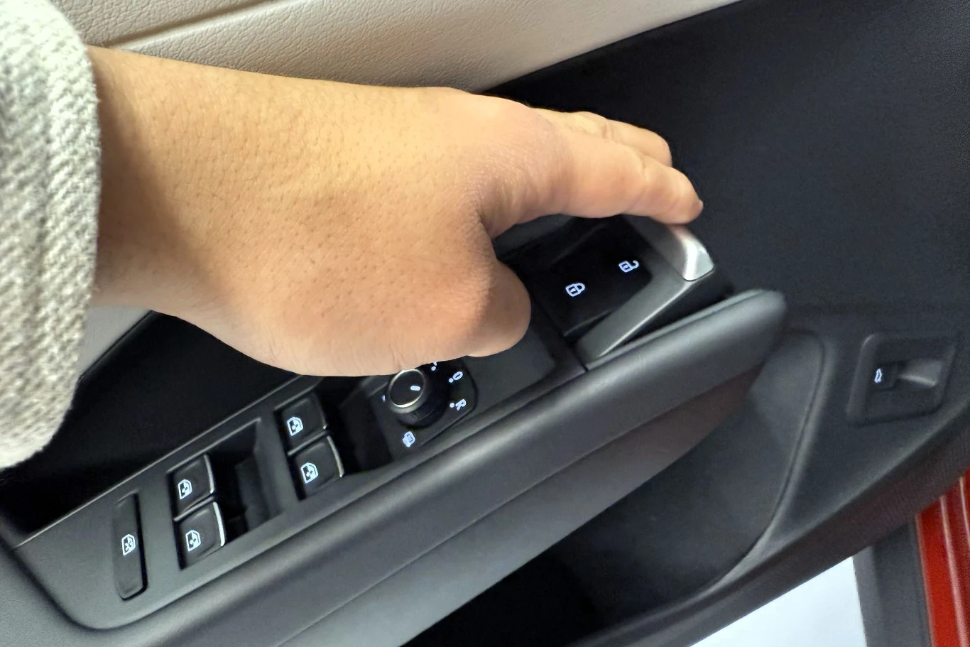 Ręka osoby naciskająca jeden z przycisków na panelu sterowania szybami i zamkami drzwi samochodu, we wnętrzu pojazdu widoczne są także inne przyciski oraz część tapicerki.