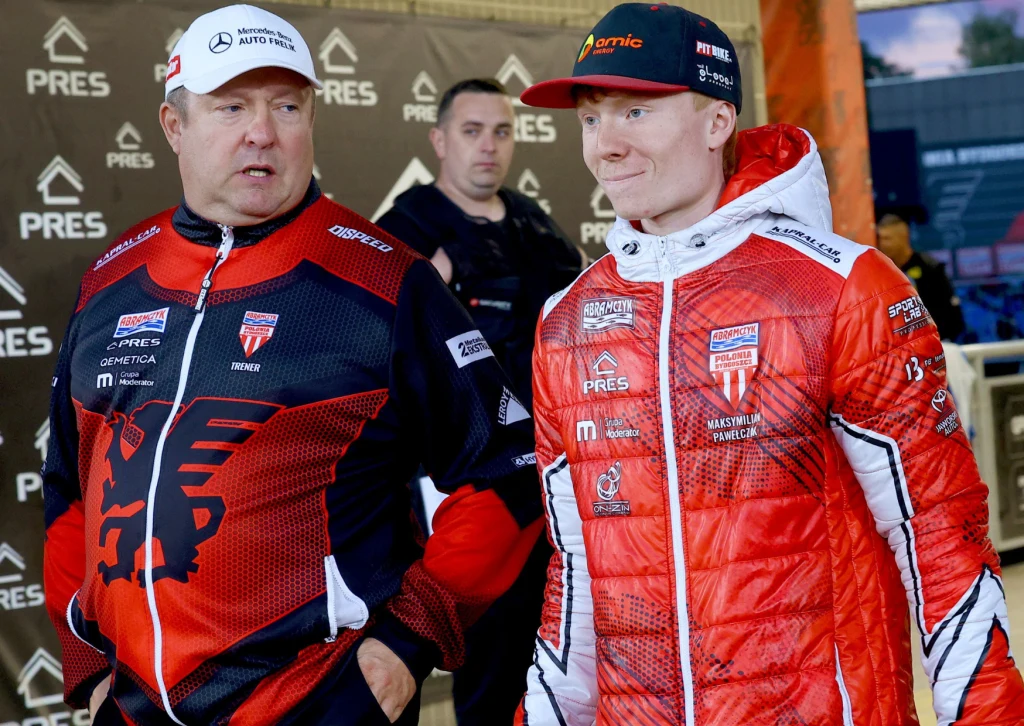 Tomasz Bajerski przed ostatnim meczem w charakterze trenera Polonii Bydgoszcz. Dwóch mężczyzn ubranych w charakterystyczne czerwone kurtki sportowe z logotypami sponsorów, stojących obok siebie; w tle baner z powtarzającym się logo PRES i inny mężczyzna obserwujący scenę.