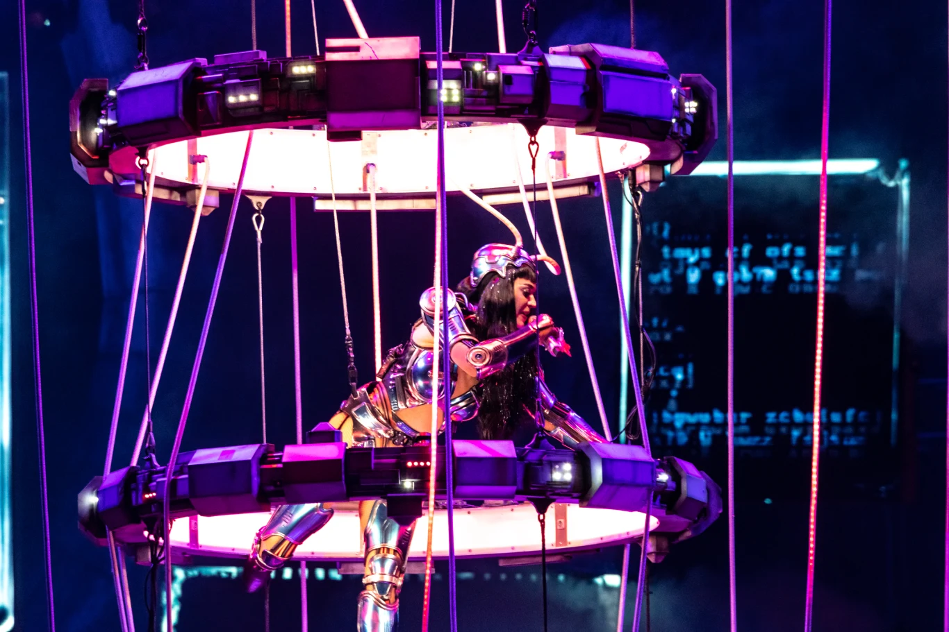 Artystka ubrana w futurystyczny, metaliczny kostium wykonuje widowiskowy występ na zawieszonej konstrukcji otoczonej światłami, tło lekko rozmazane z efektami świetlnymi dodającymi kosmicznego klimatu.