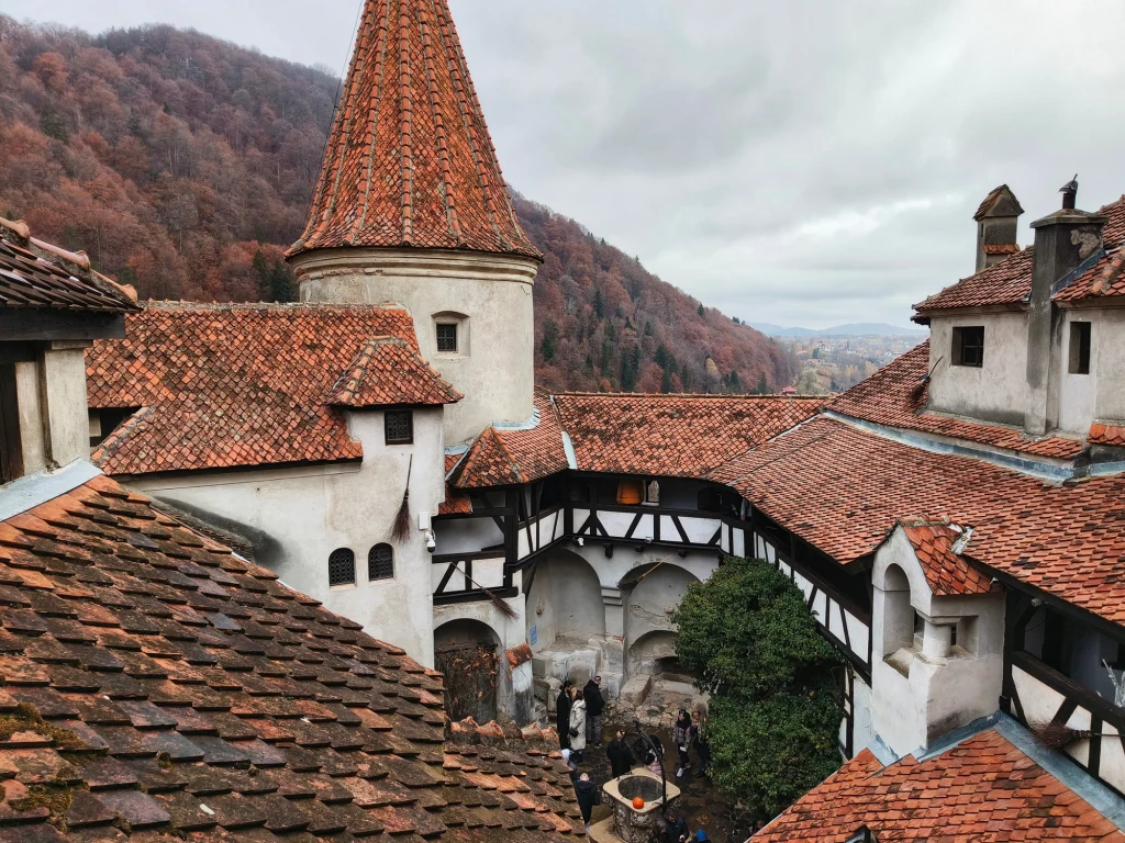 Czerwone dachówki kamiennego zamku otoczonego wzgórzami porośniętymi jesiennymi drzewami, wewnętrzny dziedziniec z grupą zwiedzających multifunkcyjne krużganki i wieżę.