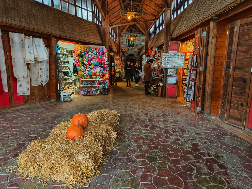 Wnętrze drewnianego budynku targowego z kolorowymi stoiskami po bokach i ozdobnymi dyniami ustawionymi na snopie siana pośrodku szerokiego przejścia, z kilkoma osobami przechodzącymi w tle.