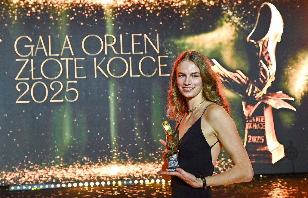 Maria Żodzik Młoda kobieta w eleganckiej sukience trzyma statuetkę, w tle widoczny napis 'Gala Orlen Złote Kolce 2025' oraz stylizowany wizerunek nagrody, scena oświetlona złotym światłem podkreślającym uroczysty charakter wydarzenia.