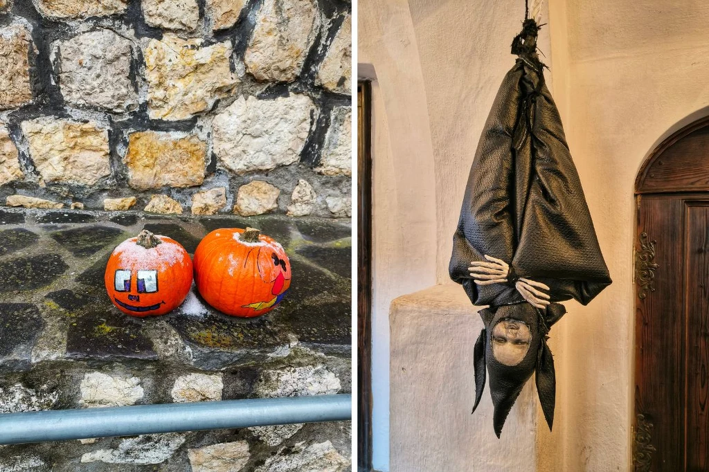 Dwie dynie ozdobione zabawnymi rysunkami ustawione na kamiennym murku oraz postać w czarnym kostiumie, zawieszona do góry nogami, stylizowana na nietoperza lub straszydło, zawieszona we wnętrzu przy ścianie.