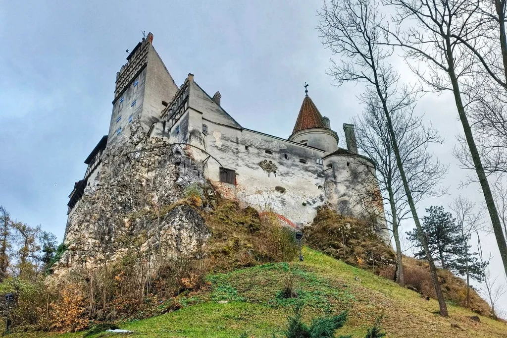 Stary zamek wznoszący się na skalistym wzgórzu, z widoczną główną wieżą, masywnymi murami i fragmentami muru obronnego, otoczony drzewami i niską roślinnością, z dominującymi odcieniami szarości i zieleni.