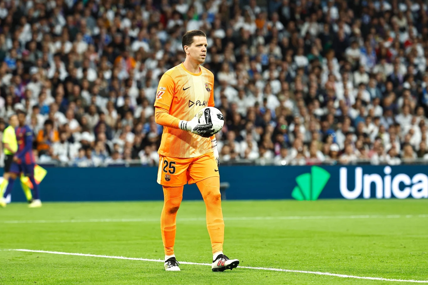 Bramkarz w pomarańczowym stroju sportowym trzyma piłkę podczas rozgrywanego meczu piłki nożnej, w tle wypełnione trybuny pełne kibiców.