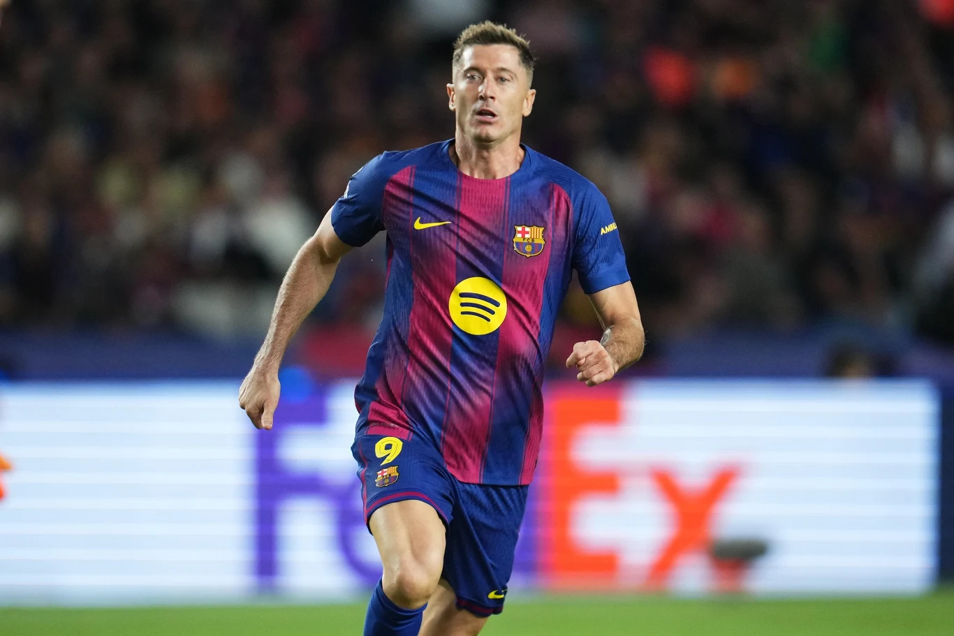 Piłkarz w stroju FC Barcelony biegnący po murawie podczas meczu, na tle rozmytego stadionu i widocznych neonowych reklam.