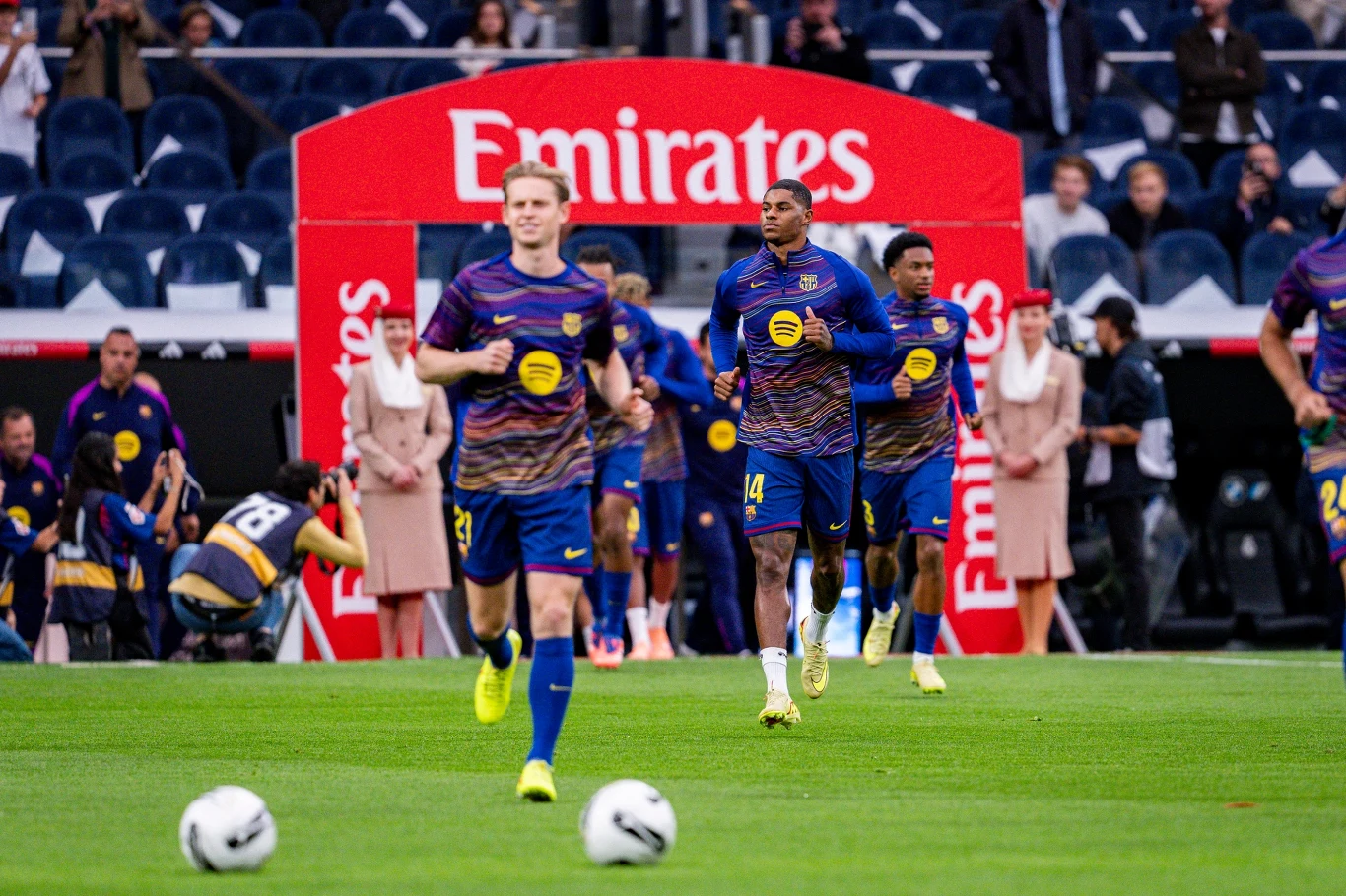 Grupa piłkarzy ubranych w niebiesko-fioletowe stroje z żółtymi elementami biegnie po murawie stadionu piłkarskiego, w tle widoczna czerwona brama sponsora Emirates oraz postacie w jasnych strojach stojące na linii bocznej.