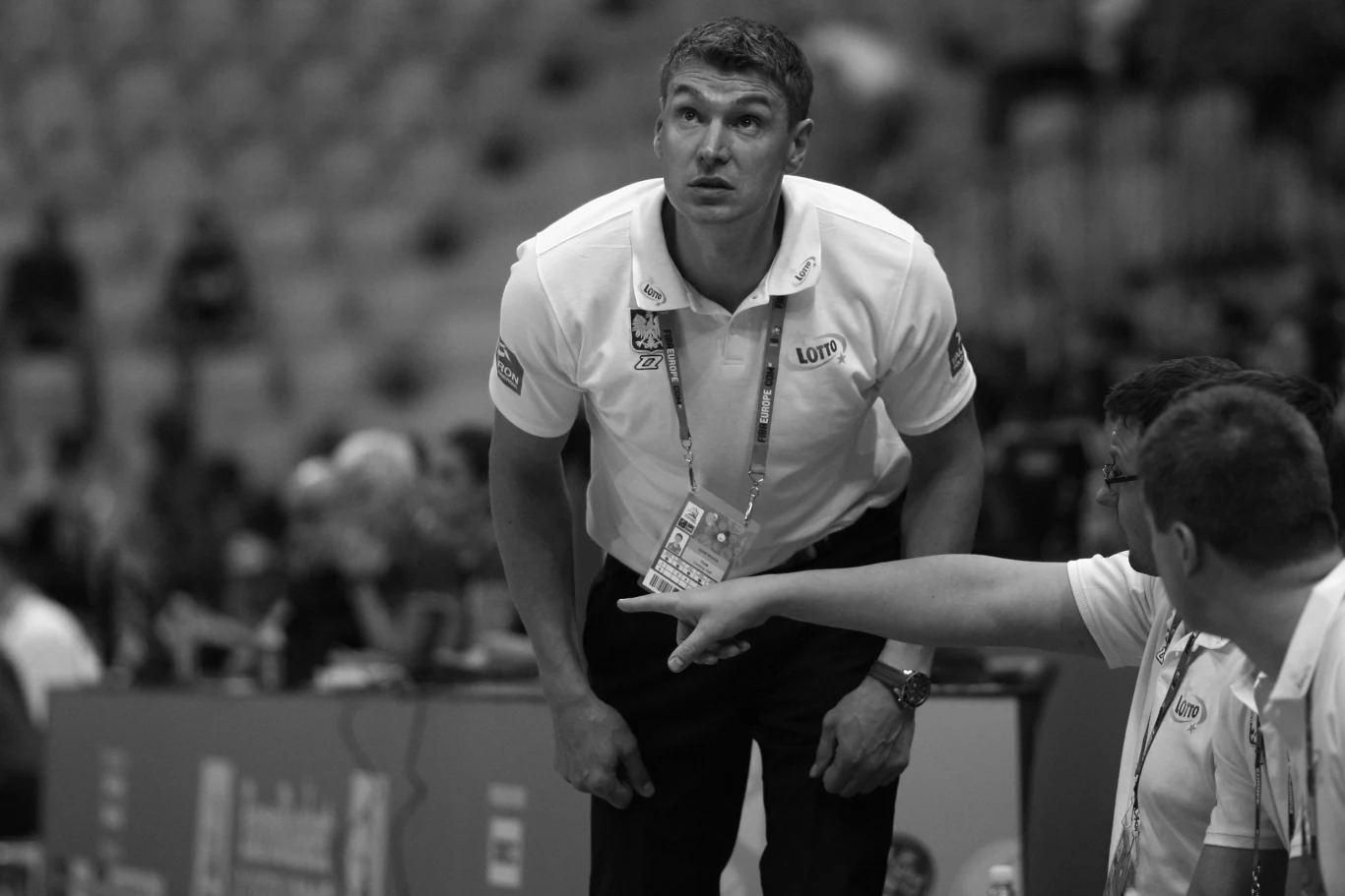 Mężczyzna ubrany w koszulkę z logotypem stoi pochylony obok siedzącej grupy osób na hali sportowej. W tle widać rozmazany tłum. Mężczyzna sprawia wrażenie skoncentrowanego i zaangażowanego w wydarzenie sportowe.