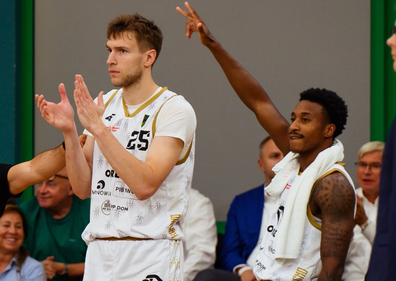 Dwóch koszykarzy w białych strojach sportowych bije brawo i gestykuluje, w tle widoczni są kibice siedzący na trybunach. Atmosfera wskazuje na sportowe wydarzenie.