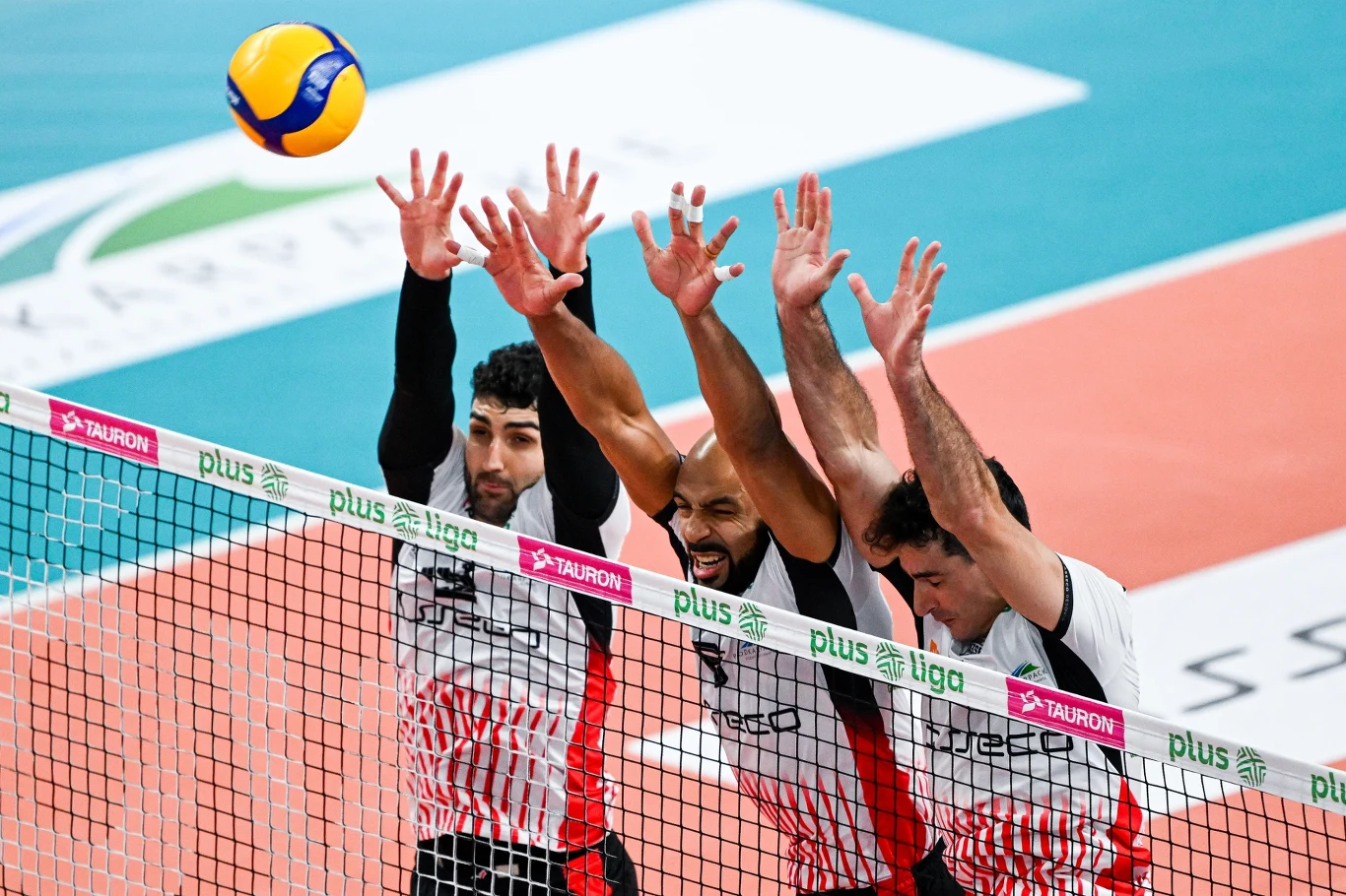 Trzech siatkarzy w biało-czerwonych koszulkach wykonuje blok przy siatce, unosząc wysoko ręce podczas meczu siatkówki, a tuż nad nimi znajduje się kolorowa piłka.