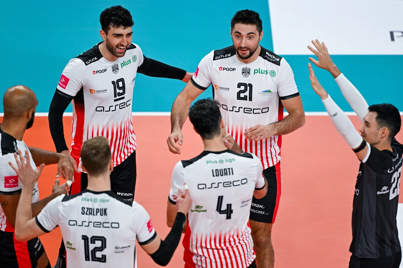Grupa siatkarzy w białych koszulkach z czerwonymi elementami na parkiecie świętuje zdobyty punkt, zawodnicy klaszczą i okazują radość, w tle fragment boiska.